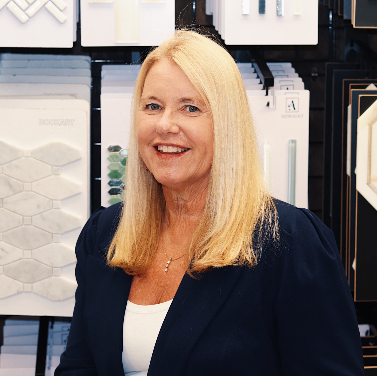 A woman with long blonde hair in a blue blazer and white shirt smiles in front of a flooring company’s geometric tile display.