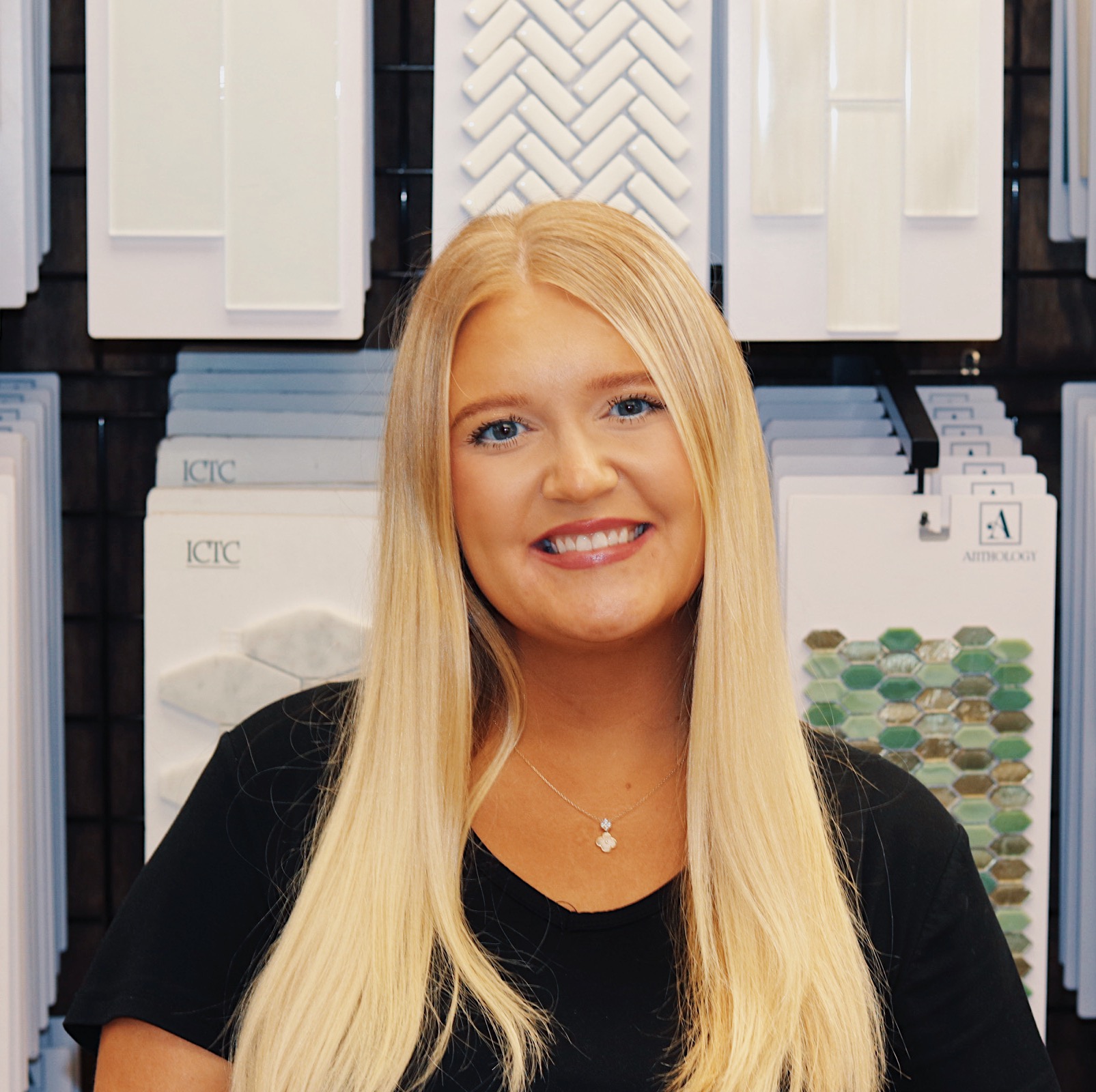 A woman with long blonde hair and a black shirt smiles in front of a flooring company wall displaying various tile samples.