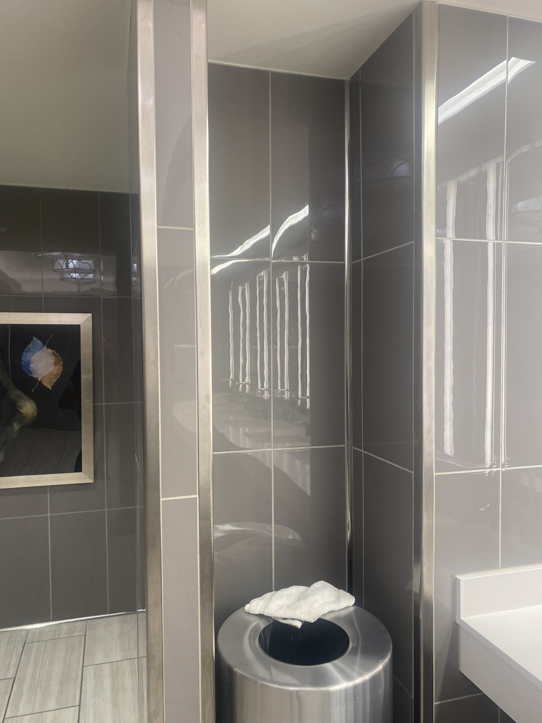 IMG_9296 A stainless steel trash can with folded paper towels on top, beside a gray tiled wall installed by a flooring company in a restroom.