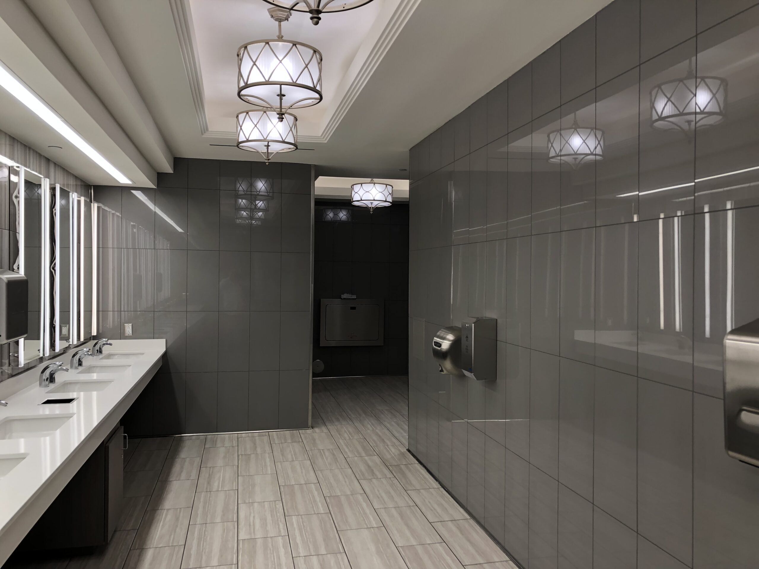 IP_7 Modern public restroom with gray tiled walls, quality flooring by a flooring company, multiple sinks, hand dryers, and a baby changing station in the back.