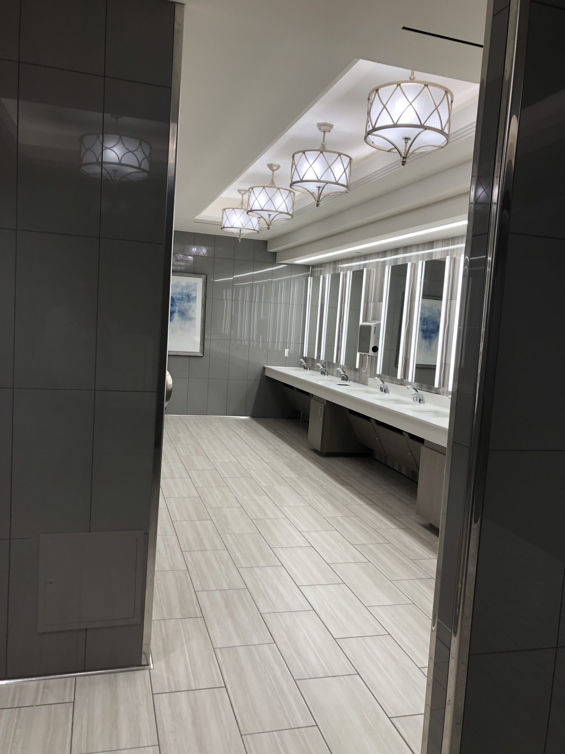 IP_9 Modern public restroom with gray tile walls, light wood flooring by a top flooring company, white sinks, large mirrors, and pendant lights.