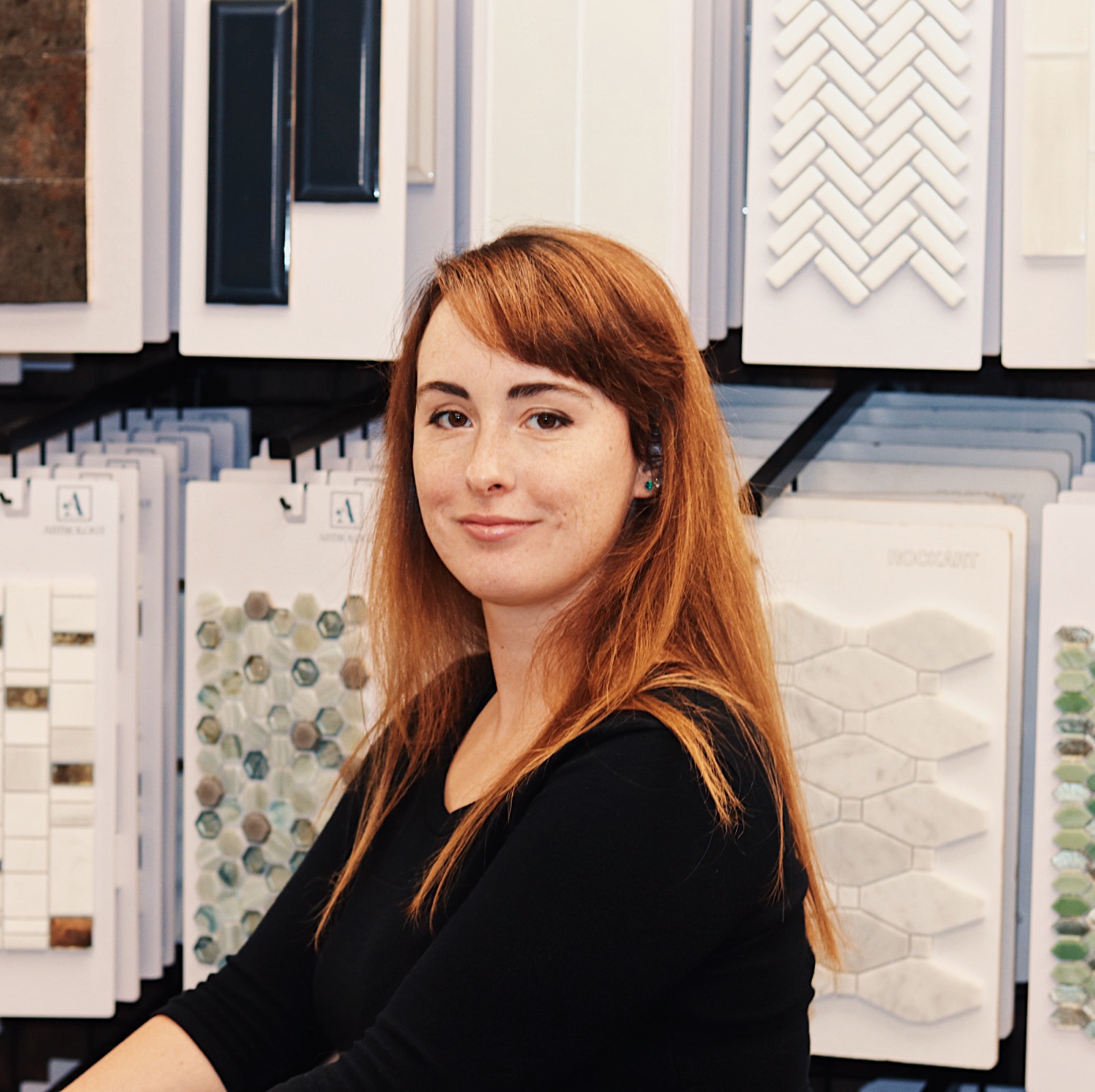 A woman with long red hair and a black top stands in front of a flooring company’s display of tile samples arranged on white boards.